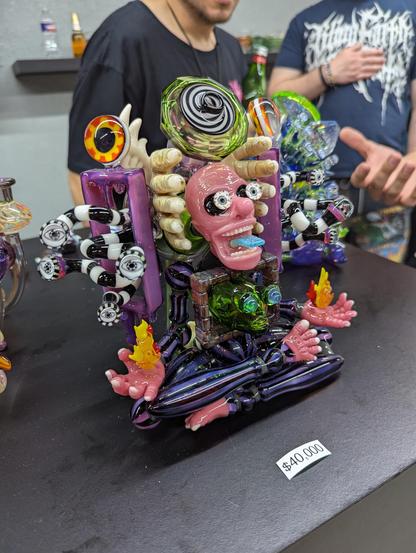 A psychedelic pipe with a cross legged human with eyes popping out and blue tongue many spiral tentacles about and swirly glowing top part. Purple and black and white glass abound on the $40000 pipe
