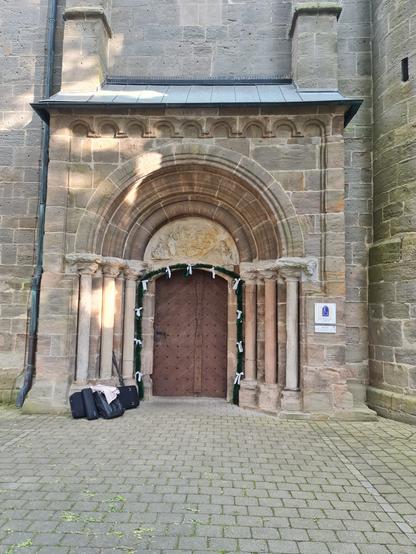 Das Bild zeigt die Eingangstür einer Kirche. Die Tür ist aus massivem Holz mit einem romanischen Bogen darüber. Der Bogen ist mit einem Relief verziert, das möglicherweise religiöse Motive darstellt. Die Tür ist mit grünen Girlanden und weißen Schleifen geschmückt, was auf eine besondere Veranstaltung hinweisen könnte. Die Tür ist von Säulen flankiert, die mit kunstvollen Kapitellen versehen sind. Vor der Tür liegen einige Koffer und Taschen,