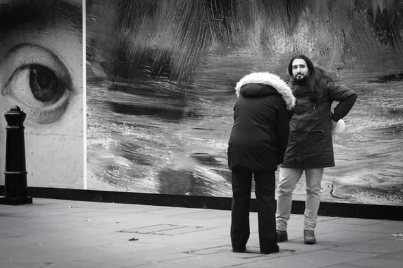 Black and white. A young couple are standing near very large photos of details from famous works of art. He is looking directly at my camera and not at the woman's phone as she takes a photo of him. At the far left of the picture, a large painted eye also seems to be looking at my camera.