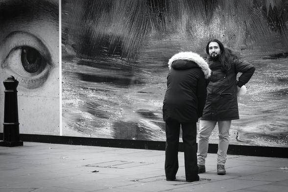 Black and white. A young couple are standing near very large photos of details from famous works of art. He is looking directly at my camera and not at the woman's phone as she takes a photo of him. At the far left of the picture, a large painted eye also seems to be looking at my camera.