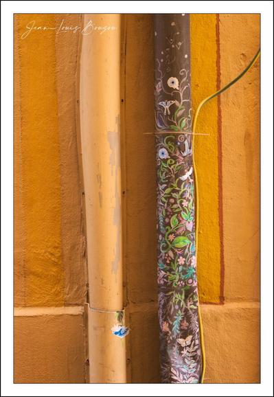 The image captures a close-up view of two adjacent vertical pipes mounted against a textured ochre-yellow wall. The pipe on the left is plain, painted in a solid beige or light tan color. In contrast, the pipe on the right is strikingly decorated with an intricate, hand-painted design featuring a muted palette of greens, blues, purples, and whites. The design showcases a natural theme, with detailed depictions of leaves, vines, and small flowers that wind their way along the length of the pipe. The background of this pipe is darker, helping the botanical patterns stand out vividly.
This scene portrays an interesting blend of functional urban elements with artistic expression, symbolizing how everyday objects can be transformed into canvases of creativity. The decorated pipe may represent efforts to beautify and personalize an otherwise mundane and industrial object, reflecting cultural tendencies to incorporate art into public or semi-public spaces. The warm yellow wall behind both pipes adds a complementary warmth to the cooler and earthy tones of the decorated pipe, enhancing the visual appeal of the composition.