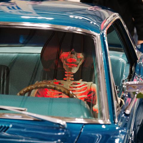 A skeleton, illuminated with red light from underneath, wearing a cowboy hat and sunglasses, is seated behind the wheel of a vintage blue car.