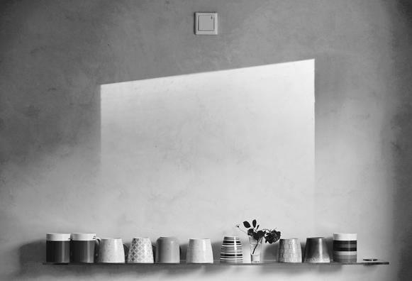Black and white photo. A row of differently shaped coffee mugs on a glass shelf at the lower border of the picture. Above that, the slightly textured wall shows a light rectangle with a slanted top edge that is made from the light coming from outside the picture. At the very top center there is a covered power outlet. The composition is very symmetrical and geometric.