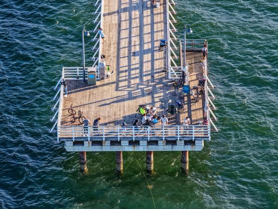 An aerial shot of the end of the Shorncliffe pier, surrounded by green and blue water.  The pier has lots of people on it fishing