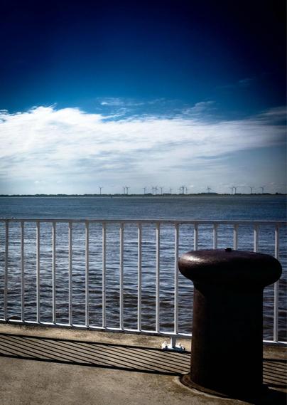 This photo captures a peaceful waterfront scene. In the foreground, there is a white metal railing and a large, dark bollard typically used for mooring boats. The surface of the water is calm, stretching across to the opposite shoreline. In the background, there are numerous wind turbines lined up along the horizon, indicating a wind farm. Above, the sky is partly cloudy with a vibrant gradient of blue, creating a serene and slightly dramatic atmosphere. Shadows from the railing fall diagonally on the concrete, adding depth to the image.