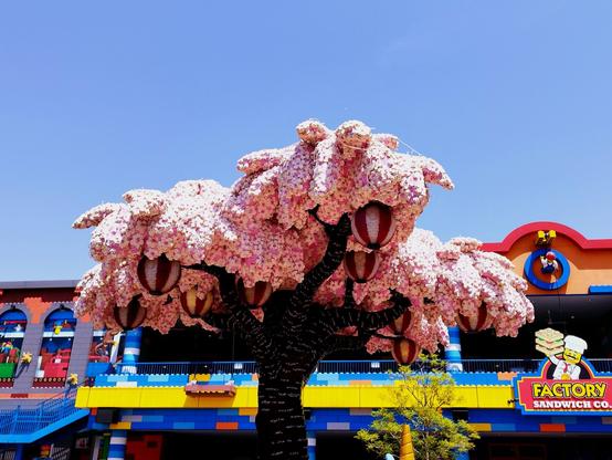 Picture of the top of a cherry tree in bloom made from Lego bricks. Buildings of the theme park in the background, sky is blue.