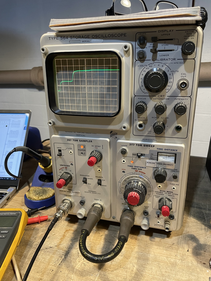 A 1960s time domain reflectometer on a bench. The instrument is silver with red and black knobs, with a CRT display showing the response of a pulse being sent into an unterminated cable and reflecting back.
