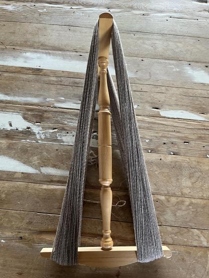 Light wooden niddy noddy with a whole about 190 grams of very thin 3-ply yarn on it. It's on a slightly darker rough wooden table with patches of white paint.
