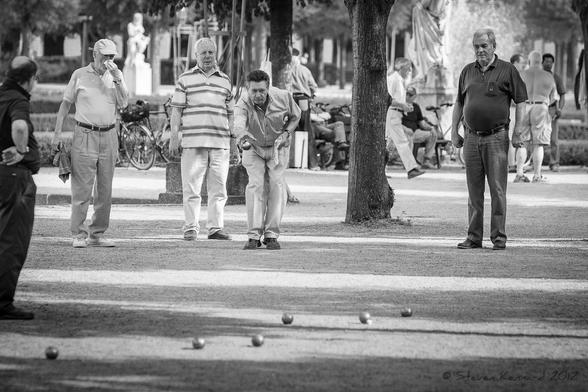 A black and white photograph of some men in a public park playing a game of boules. One man has just released the boule which is flying from his hand, while the others look on. There are others scattered on the foreground. Behind the men are trees and bicycles and people enjoying the park.