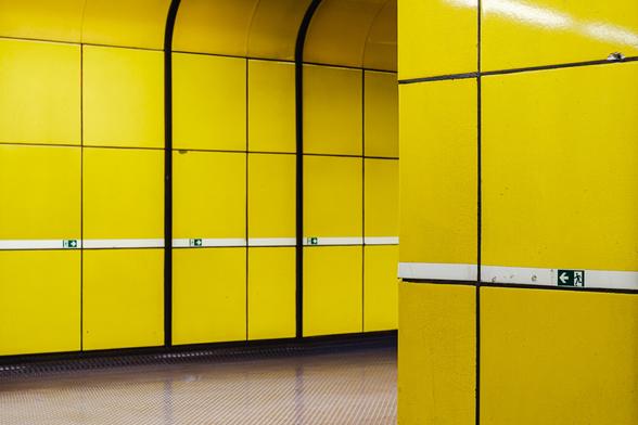 Photo of a brightly lit yellow subway or metro corridor with smooth tiled walls and black dividing lines. Small green emergency exit signs are mounted along a horizontal white stripe running through the middle of the tiles.