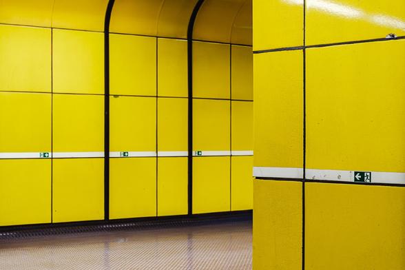 Photo of a brightly lit yellow subway or metro corridor with smooth tiled walls and black dividing lines. Small green emergency exit signs are mounted along a horizontal white stripe running through the middle of the tiles.