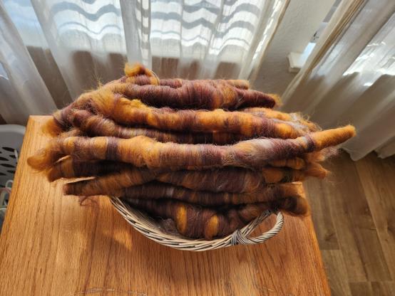 A basket sits on a wooden table near a curtained window. The basket is full of rolags, which are long, sausage shaped rolls of multi-colored wool, like very tight hair curls. The rolags are orange, brown, and grey.