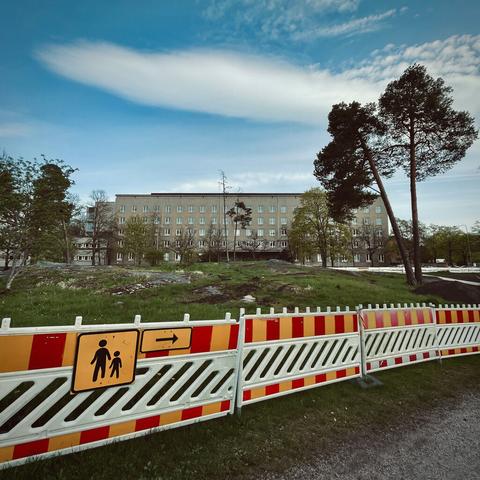 Construction barriers with a pedestrian sign in front of a large, rectangular multi-story building under a blue sky. Two pines can be seen at the right side of the photo.