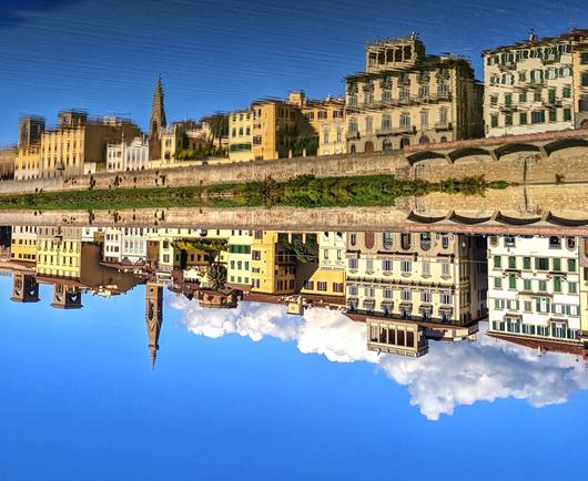 An upside-down view shows a row of buildings along a riverbank, reflected in the water. Above the reflection is a bright blue sky with fluffy white clouds. The buildings are a mix of pale yellow and cream colours, with various window styles. A tower with a spire is visible amongst the buildings. The river appears calm with slight ripples causing distortions in the reflections.