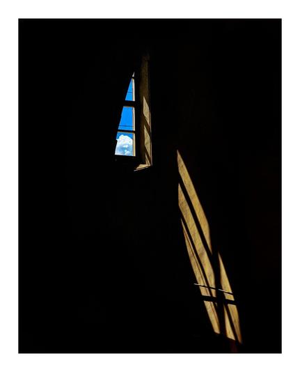 The photo shows a small, triangular window in a dark room.  Through the window you can see a patch of bright blue sky with a fluffy white cloud in the bottom pane. Strong sunlight casts shadows of the window panels on the dark wall, creating a dramatic contrast between light and shadow.
