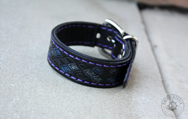 A black leather bracelet with dragonscale stamping and silver accent scales. It has a slim black liner, purple stitching, and nickel plated brass hardware.