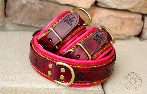 A handmade leather collar and pair of cuffs. They have burgundy top straps with custom dragonfly stamping over mahogany base straps and bright pink liners. They have yellow stitching and solid brass hardware.