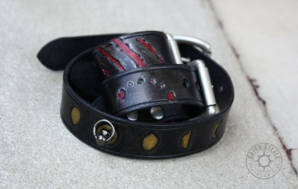 A handmade leather day collar and pair of cuffs. They're solid black leather with a slight metallic gray shimmer and nickel plated brass hardware. The collar has the phases of the moon tooled and painted metallic gold, and the cuffs have tooled and painted clawmarks and bitemarks.