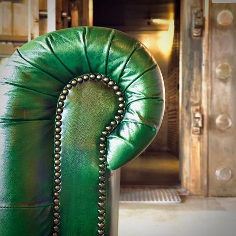 The left arm of a deep green, modern leather-upholstered, button tufted, chesterfield-styled sofa fills frame left, with a bank vault's open door in the distant frame right.