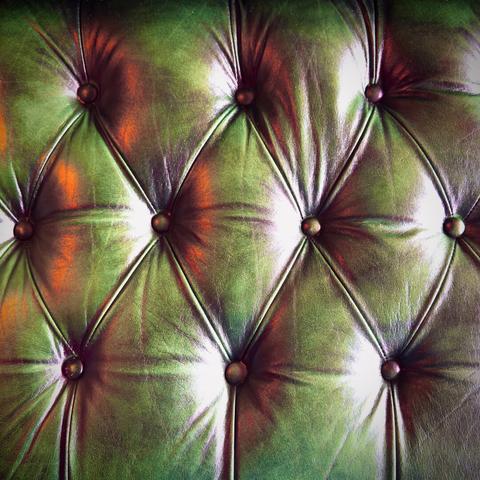The back of a deep green, modern leather-upholstered, button tufted, chesterfield-styled sofa fills the frame under amber accented lighting.