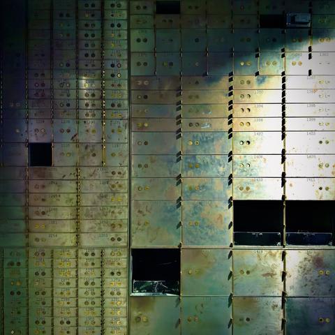 A patina-colored wall of mostly closed safe deposit boxes (with a few open and pitch-black on the inside) reflects light shining from out of frame right.