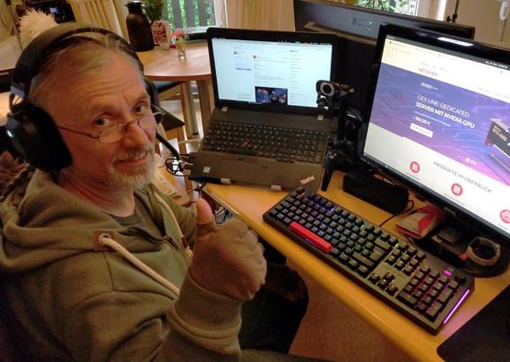 Der Gewinner vom Hetzner Giveaway sitzt mit neuen Corsair Gaming HS80 RGB Headset und neuer Tastaur sitzt am Tisch vor einem Bildschirm und einem Laptop. Er lächelt in die Kamera und hält den Daumen hoch.