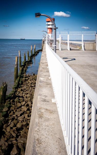 This photo shows Pier 4 near Brunsbüttel on the Elbe. In the foreground, a sturdy white metal railing and a wide pedestrian walkway are visible. To the left of the pier, a rocky stretch of shoreline with old, moss-covered wooden posts stretches out.

At the end of the pier, a distinctive red and white lighthouse can be seen. Several street lamps are located along the pier. The sky is clear with only a few small clouds, the water calm, and a ship can be seen on the horizon. The overall scene is bright and peaceful, with a strong sense of texture and a maritime atmosphere.