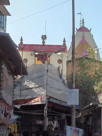 The image appears to depict a temple entrance, likely in India, marked by a sign that reads "श्री महालक्ष्मी मंदिर" (Shri Mahalaxmi Mandir). There is a visible timer or clock displaying the number "54." The structure seems to be under a canopy, with additional temple architecture and decorations visible in the background. The surrounding area appears to be bustling with people.

Image Credits: Wikimedia / No machine-readable author provided. Nichalp assumed (based on copyright claims). / CC BY 2.5