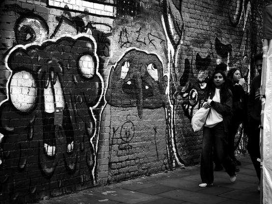 Black and white. In Brick Lane Market, a woman and a man, on the far right of the picture, are walking to the left from behind one of the stalls, past a wall with three pieces of street art. The piece on the left and right are almost unrecognisable but probably represent monsters of some kind. The one in the middle resembles a bald-headed man with a long droopy pointed nose that reaches down past a mouth with only two teeth. His eyes both point outwards. The effect is of a a person who is exasperated by being photobombed while having his picture taken by me.