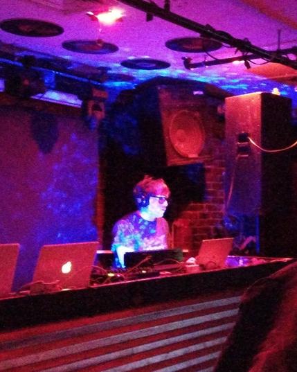 A man with short hair and glasses, wearing headphones, while DJ'ing in front of a computer and DJ equipment. There are red and blue and purple lights.