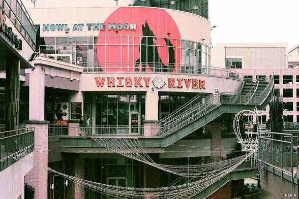 Farbfoto - Blick auf einen Teil eines Einkaufszentrums in Charlotte, North Carolina, USA. In Bildmitte eine zum Zeitpunkt der Aufnahme geschlossene Bar mit dem Namen "Whisky River". Darüber das Wandbild eines heulenden Wolfs und die Beschriftung "How at the moon". Moderne sachliche Architektur mit Treppen und Geländern. Leica R4 mit Summilux 50 mm auf Lomochrome Purple-Film.