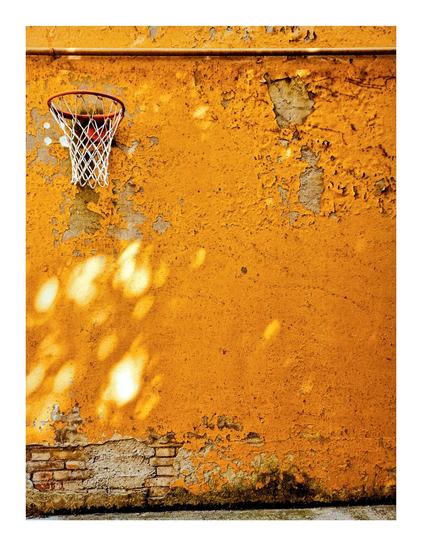 The photo is of a weathered, yellow/ochre-coloured wall with a basketball hoop on it on the top left.
There is a pipe at the top of the photo running across it which creates a sort of natural frame. The paint on the wall is heavily chipped and flaking, revealing the underlying brickwork at the base and near the top right under the pipe. The sunlight casts shadows on the surface of the wall under the basketball hoop.
