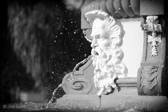 This Is A Black And White Photo In Landscape Format. To The Right Of Centre Is The Side Of A Small City Fountain Located In A Park. On The Side Is The Sculpture (Probably A Metal Casting) Of A Man's Face Painted Brilliant White And Viewed Nearly In Profile. From The Mouth Is An Unsteady Stream Of Water Descending Into The Base Of The Fountain. The Flow Is Frozen By A Fast Camera Shutter. Surrounding The Face Are Multiple Droplets Of Water Being Thrown Into The Air By The Stream Of Water. The White Face Is Framed By Ornate Scrolls In A Very Light Grey. Behind Is A Dark Background Of Trees, All Heavily Blurred Out Of Focus And In Shadow. 
The Location Is The Esplanade Park In Singapore. The Fountain Is The Tan Kim Seng Fountain Erected In 1882 In Honour Of The Man Of The Same Name (2012).