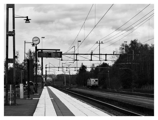 En liggande svartvit bild på en liten järnvägsstation, ingen byggnad, bara en perrong och ett par spår. 2 vita, breda streck leder bort på perrongen och till höger står ett lok. Till vänster stolpar, en skylt hänger och visar avgångar. Ovanför den är en klocka som visar 1445. Himmelen är full av moln med strukturer.