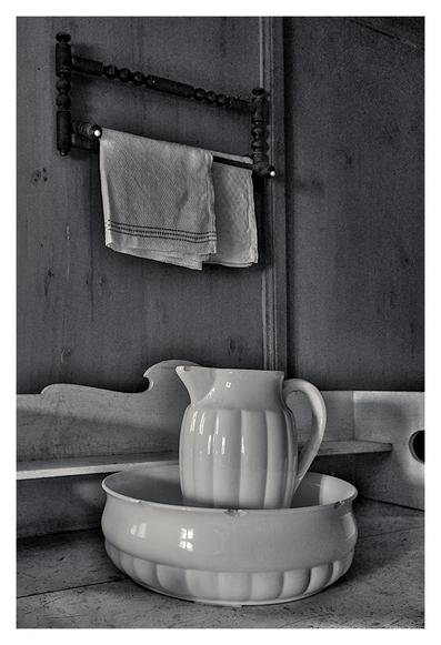 The picture shows a black and white image of a rustic scene. In the foreground is a large, white wash bowl with a wavy rim on a simple, wooden washstand. In the bowl stands a matching wash jug with a handle and a spout, also made of white ceramic. Both have minor damage to the edges. In the background, a towel hangs from a wooden towel rail attached to a rough wooden wall. The lighting is soft, with light coming in from the left and casting gentle shadows. The scene conveys a sense of simplicity and nostalgia.