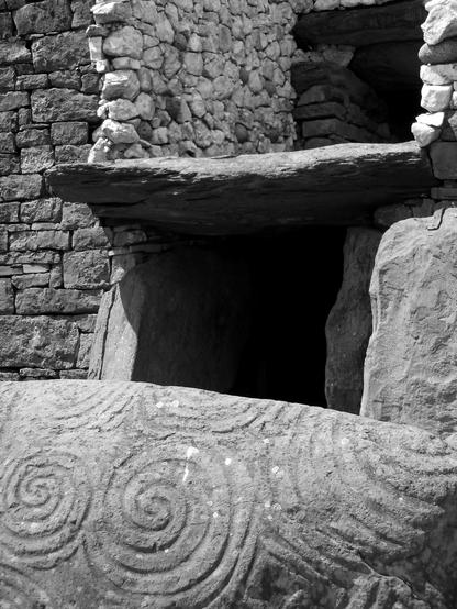 Black and white photo of the entrance to a monumental prehistoric tomb. Large boulders - topped by a single stone slab - flank the darkened entranceway at ground level. Just above that, is a deep recess which is faced with light colored, irregularly shaped stones that contrast with the darker, rough ashlar masonry of the tomb’s exterior. A wide boulder - featuring incised geometric patterns - occupies the foreground.