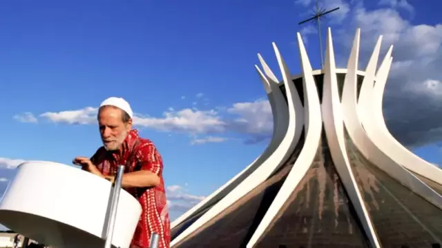 Sergio Boré, an older man in a red shirt and white cap, playing a large steel drum in front of a futuristic buidling against a blue sky with few clouds.