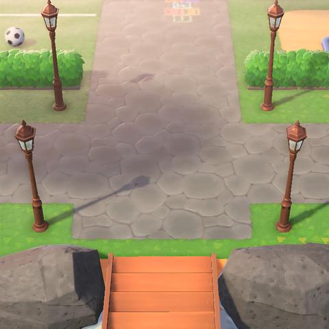 Animal Crossing: New Horizons screenshot. It's set at the entrance to the island. The entrance which has a concrete walkway in front of them in all cardinal directions. On either side of the entrance are copper colored streetlamps. Another set of them can be seen north at an entrance to a park-like area with hedge fences. To the left a soccer ball can be seen beyond the hedges and to the right is a sandy area beyond the hedges.