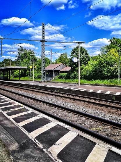Bahnsteig und Gleise im Frühling. Eine Bahnuhr zeigt 12 Uhr.