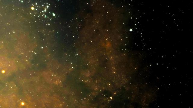 A bright star cluster in the top left sits in front of a cloudy nebula that fades to the right. In the upper right is a second, fainter cluster. There is a pair of orange stars on either side of the bottom left corner.