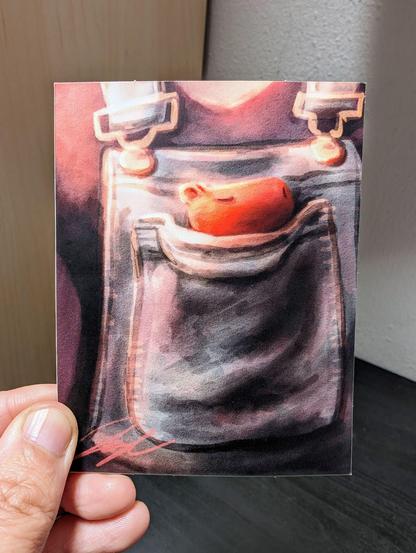 Photo of a left hand holding a rectangle sticker. The sticker is a matte painting of a capybara snuggling in a pocket.
