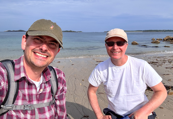 Two men on a beach. They look to camera. One is wearing checkered shirt and green cap. To his right is a man in white t-shirt and beige cap. He wears sunglasses. Behind is the sea and distant islands