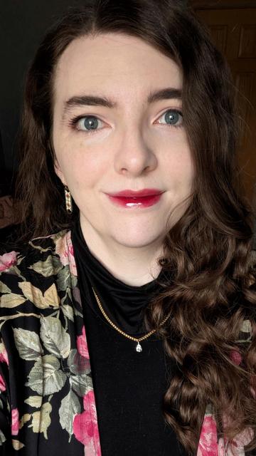 selfie of Tulip smiling. they're wearing a pink and black floral shawl, a black turtleneck, and a golden necklace on top. they're also wearing a glossy deep pink lipgloss