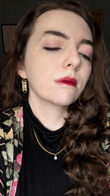 Tulip looking down and away from the camera, highlighting the wash of light pink eyeshadow on their eyelids. they have pale skin, dark eyebrows, and long dark brown waves framing their face