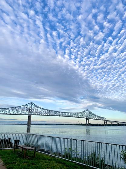A view of the blue sky, scattered with many patches of clouds, stretches above the long cantilever bridge that spans the wide Delaware River. A grassy area lies in the foreground.