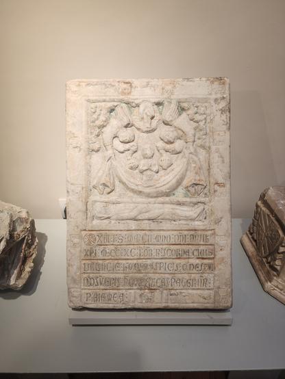rectangular grave stone with 4.5 lines of abbreviated Latin text in bottom half and a carved scene in top half. the scene shows the deceased person lying in the grave at bottom and, above him, his soul in a canopy being lifted up to heaven by angels who are also dangling censers.