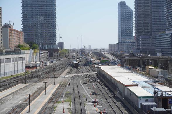 A photo looking down along a rail corridor just beyond a large station. The photo is taken from a couple of storeys up. Eight tracks are visible in the foreground, as well as ends of four train platforms. There are many crossover tracks and the corridor narrows to six tracks a bit further away from the camera.

A train of five passenger cars behind a locomotive is just departing and is in an S shape as it goes through two crossovers around something that looks like a small infrastructure box. A bit further in the background is a longer oncoming train that looks to be heading straight for where the rear cars of the first train are; it will turn off to the side on a track curve that isn't visible because the first train is blocking the view.

None of the tracks are electrified. An overhead signal gantry immediately beyond the platform has signals blocked off with white "X" shapes, and signal heads are at ground level instead. There is another large signal gantry further down.

Immediately to the right of the corridor is a construction zone with temporary portable buildings and a work area, and beyond that is an elevated highway with ramps up and down to street level. There are high-rise residential buildings on both sides. The middle of the photo has a more distant horizon and two tall smokestacks, an overhead electricity transmission line, and a few construction cranes are visible.

It is sunny; the sky looks blue-white-grey and slightly hazy, maybe a bit smoggy.