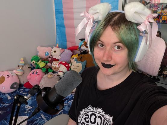 Revy looking up at you
She's wearing black lipstick and cat ear headphones and is near a microphone 
Many plushies in background and trans flag