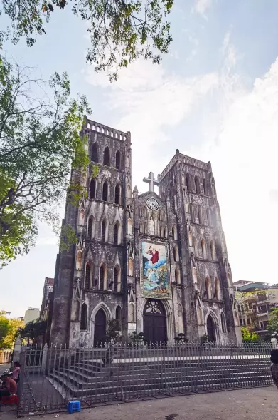 The image depicts a large cathedral or church, featuring Gothic architectural elements such as tall towers, pointed arches, and ornate details. There is a prominent cross at the top, and a large painting or mural visible on the facade. Surrounding the building are trees and possibly a fence or gate in the foreground. The overall atmosphere suggests a historic and significant structure, likely in an urban setting.

Image Credits: Wikimedia / Arno Kohlem / CC BY-SA 4.0