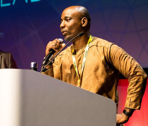 Waist-up color photo of Wole Talabi, a Black man with a shaved scalp and a short salt-and-pepper beard. He wears a tan-colored woven shirt with yellow detailing at collar, yoke, and cuffs; a lanyard around his neck; and a black fitness tracker on one wrist. He is standing in relaxed posture, speaking into a microphone on a podium that takes up about a quarter of the rectangular image. He appears alert but comfortable.